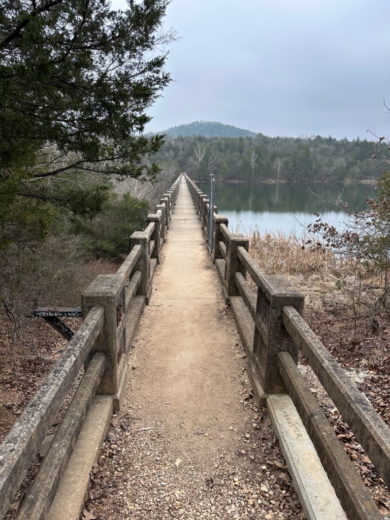 A long, narrow walkway leading across a calm body of water, surrounded by trees and mountains in the background on a cloudy day.